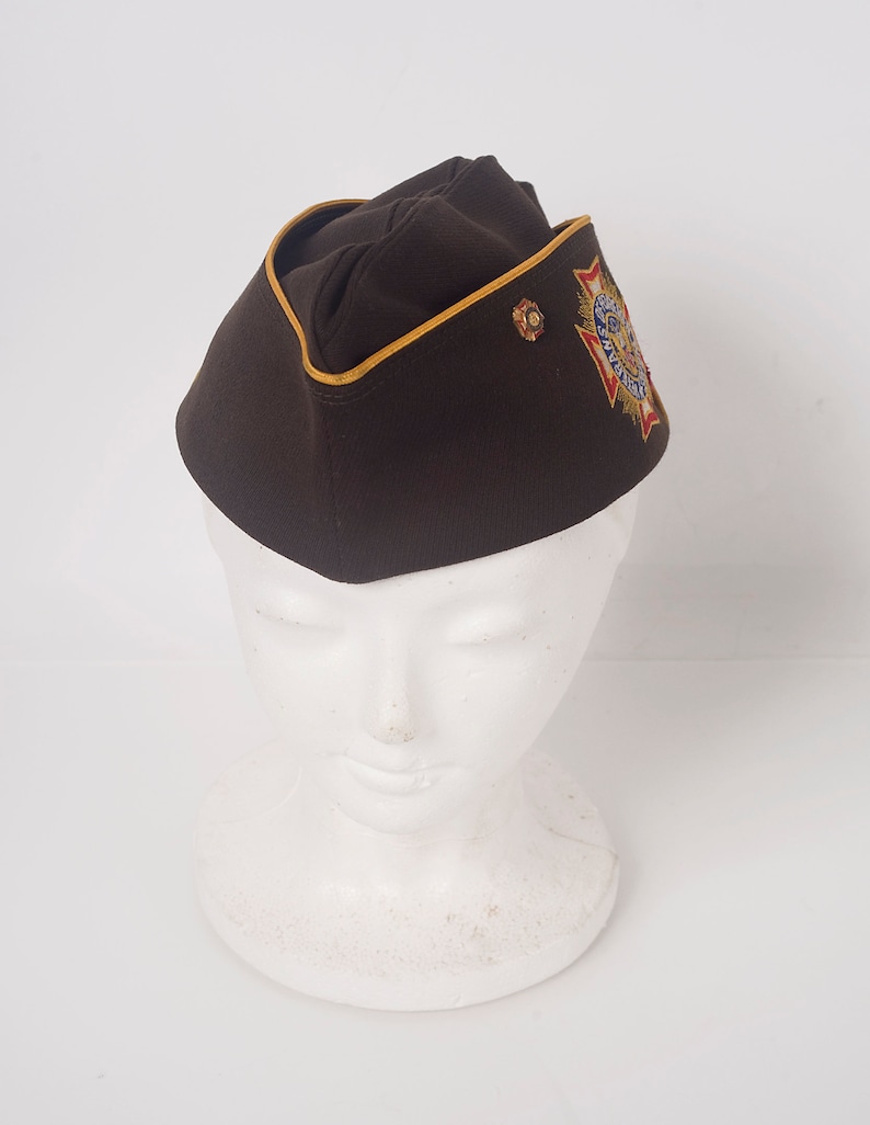 May include: A brown military-style cap with a gold trim and an embroidered patch featuring a red, white, and blue star with the words "American Legion" around it.