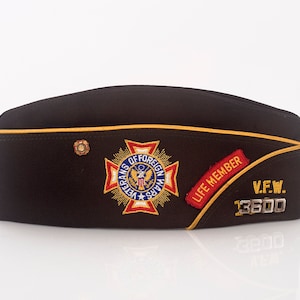 May include: A brown felt military-style hat with a gold trim and a red, white, and blue embroidered patch that says "Veterans of Foreign Wars". The hat also has a red and gold patch that says "Life Member" and a silver badge that says "V.F.W. 13600".