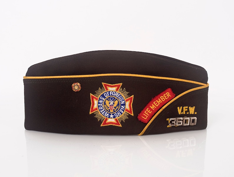 May include: A brown felt military-style hat with a gold trim and a red, white, and blue embroidered patch that says "Veterans of Foreign Wars". The hat also has a red and gold patch that says "Life Member" and a silver badge that says "V.F.W. 13600".