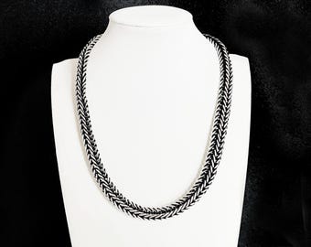 Puede incluir: Un collar negro y plateado con un diseño de espiga se exhibe en un maniquí blanco. El collar está hecho de piezas de metal entrelazadas, creando una apariencia texturizada y tejida. El fondo es negro liso.