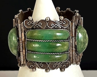 May include: A wide, ornate bracelet with a silver-colored metal frame. The bracelet features large, oval-shaped green stones on the sides and three rectangular green stones in the center. The metalwork includes decorative swirls and filigree.
