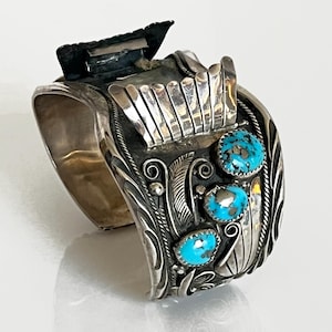 May include: A wide, silver cuff bracelet with intricate detailing and three turquoise stones. The bracelet features a decorative design with feathers and other embellishments. The cuff has a rectangular piece at the top.