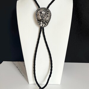 May include: Silver bolo tie with a detailed oval pendant featuring an ornate design. The pendant is attached to a black braided cord that ends with silver tips. The bolo tie is displayed on a white mannequin.