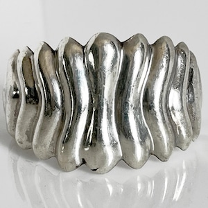 Op de afbeelding: Een zilverkleurige ring met een golvend, getextureerd ontwerp. De ring heeft een reeks verticale, langwerpige rondingen die een uniek, opvallend patroon creëren. De ring is waarschijnlijk gemaakt van metaal en heeft een gepolijste afwerking.