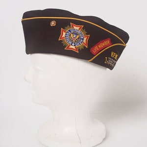 May include: A brown felt military-style hat with a gold trim and a patch that reads "Veterans of Foreign Wars" with a red, white, and blue American flag. The hat also has a red patch that reads "Life Member" and a silver pin that reads "VFW 13500".