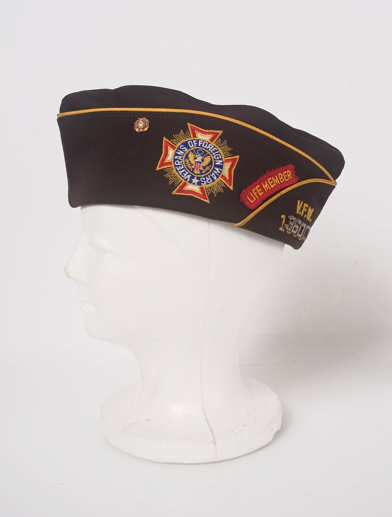 May include: A brown felt military-style hat with a gold trim and a patch that reads "Veterans of Foreign Wars" with a red, white, and blue American flag. The hat also has a red patch that reads "Life Member" and a silver pin that reads "VFW 13500".