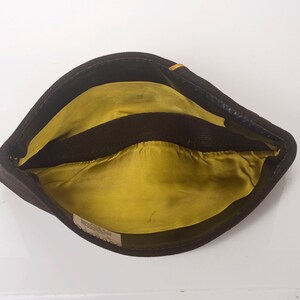 May include: A brown felt hat with a yellow lining and a black band. The hat has a small label on the inside.