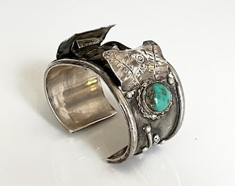 Puede incluir: Un brazalete de plata con una piedra turquesa y detalles decorativos de hojas y plumas. El brazalete abierto tiene una banda ancha y adornos detallados, mostrando un diseño del suroeste o nativo americano.