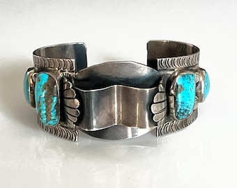 May include: A silver cuff bracelet with three rectangular turquoise stones. The bracelet has a central shield-shaped design and decorative edges. The turquoise stones have brown and blue patterns.