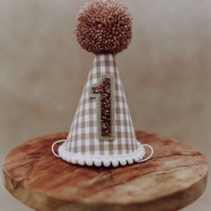 Puede incluir: Un gorro de fiesta a cuadros beige y blanco, coronado con un pompón marrón. El número "1" está aplicado con purpurina. El gorro tiene un ribete de pompón blanco y cuerdas elásticas blancas.