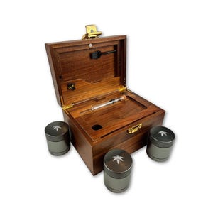 Puede incluir: Una caja de madera con tapa con bisagras, un cierre dorado y una cerradura pequeña. Dentro de la caja hay un tubo de vidrio y una herramienta negra. Tres amoladoras cilíndricas grises con un diseño de hoja en la parte superior están frente a la caja.