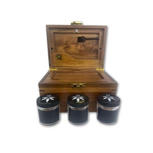 May include: A wooden box with an open lid, revealing a compartment with a small tool. Three black containers with silver rims and white floral designs are in front of the box. The box and containers are on a white background.