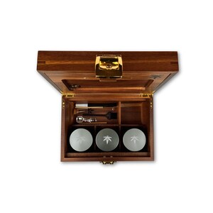 Könnte beinhalten: Eine rechteckige Aufbewahrungsbox aus Holz mit einem goldfarbenen Metallverschluss, die geöffnet ist. Das Innere enthält drei runde silberne Behälter mit Blattsymbolen, eine kleine Bürste, ein Metallwerkzeug und einen dünnen Stab in Fächern.