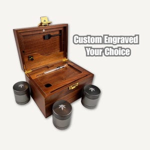 Puede incluir: Una caja de madera con tapa con bisagras, abierta para revelar un compartimento con un tubo de vidrio y un cepillo pequeño. Tres molinillos cilíndricos con un diseño de hoja están dispuestos alrededor de la caja. El texto "Custom Engraved Your Choice" es visible.