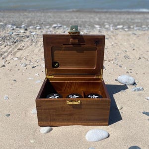 Puede incluir: Una caja de madera marrón con tapa con bisagras, abierta para revelar tres recipientes circulares negros con diseños blancos. La caja tiene herrajes de latón y está sobre una playa arenosa. La tapa está asegurada con un cierre verde.