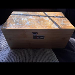May include: A rectangular wooden storage box featuring a light brown finish and a white laser-engraved tropical leaf pattern on the lid. The front of the box includes a silver-coloured metal combination lock mechanism with three rotating number dials.