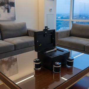 May include: A white ceramic mug with a black handle and the text "But First, Coffee" in a bold, black font. The mug is set against a blurred background, suggesting a cosy setting.