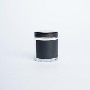 May include: A clear glass jar with a cylindrical shape, featuring a black screw-top lid and a rectangular black label centred on the front. The container is empty and has a smooth, transparent surface, providing a neutral and functional storage vessel.