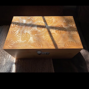 May include: A light brown wooden box with a carved tropical leaf pattern on the lid. The box has a combination lock. The box is rectangular and sits on a textured surface.