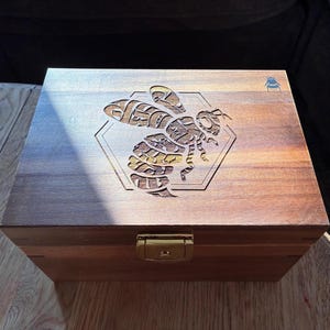 May include: A rectangular wooden box with a hinged lid featuring a laser-engraved bee design centred within a hexagonal outline. The box has a polished gold-tone metal latch on the front and a small bee icon engraved in the top right corner.