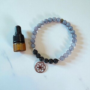 May include: A light purple beaded bracelet with a copper charm and black lava beads. A small amber glass bottle with a black dropper is next to the bracelet. The bracelet is approximately 17.8 cm in circumference.