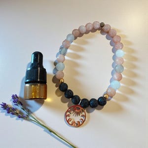 May include: A beaded bracelet with pink, blue, and black beads, and a copper-colored pendant. A small amber-colored bottle with a black dropper and a sprig of lavender are also in the image.