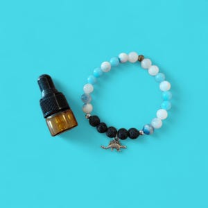 Könnte beinhalten: Ein Armband mit weißen, blauen und schwarzen Perlen und einem kleinen Dinosaurier-Anhänger. Daneben eine kleine braune Glasflasche mit schwarzem Verschluss. Der Hintergrund ist einfarbig hellblau.
