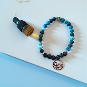 Puede incluir: Una pulsera de cuentas con un colgante de montaña y una pequeña botella de vidrio ámbar. La pulsera presenta una mezcla de cuentas azules, negras y turquesas. El colgante es un diseño circular con una silueta de montaña y sol.