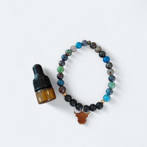 Pode incluir: Uma pulseira de contas com um pingente de touro de bronze e um pequeno frasco âmbar de óleo essencial com tampa preta. A pulseira apresenta uma mistura de contas coloridas, incluindo azul e verde.