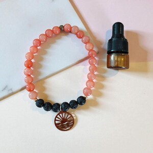以下が含まれることがあります： ピーチ色の石と黒溶岩ビーズのビーズブレスレット。太陽と波のデザインの銅色のチャームがブレスレットからぶら下がっています。黒いスポイトが付いた小さな茶色のガラス瓶がブレスレットの隣にあります。