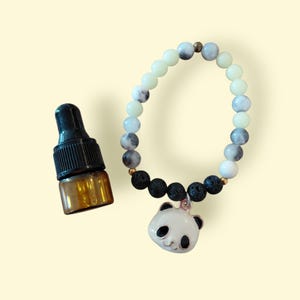 Könnte beinhalten: Ein Perlenarmband mit einem Panda-Anhänger und einer kleinen braunen ätherischen Ölflasche. Das Armband hat weiße, graue und schwarze Perlen. Der Panda-Anhänger ist weiß mit schwarzen Akzenten.