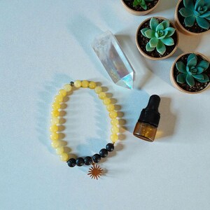 Pode incluir: Uma pulseira de contas amarelas com contas pretas e um pingente de sol dourado. Uma ponta de cristal transparente, pequenas suculentas em vasos e uma garrafa de vidro marrom com conta-gotas também são visíveis.