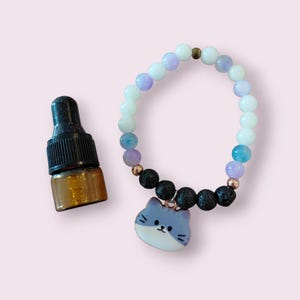 May include: A bracelet featuring white, lilac, and blue beads, black lava beads, and a blue cat charm. Beside the bracelet is a small brown glass bottle with a black dropper top.