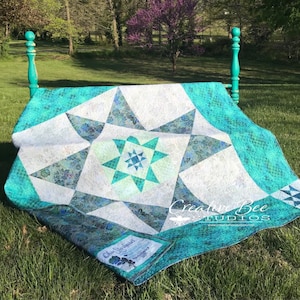 May include: A teal and white quilt with a star pattern is displayed on a teal painted bed frame. The quilt is made with a variety of fabrics and has a label that reads "Chris Stoddert Designs". The quilt is displayed on a bed frame in a grassy area with trees in the background. The text "Creative Bee Studios" is visible in the lower right corner of the image.