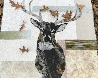 May include: A square fabric quilt featuring a central applique of a deer head silhouette crafted from camouflage patterned fabric. The background consists of various rectangular patches in shades of cream, beige, and olive green with textures.