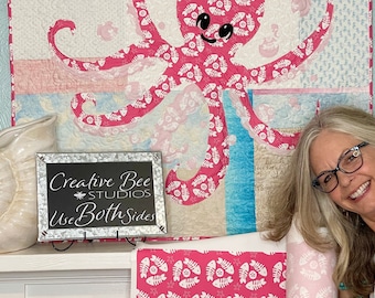 Puede incluir: Una colcha de color rosa y blanco con forma de pulpo con un patrón de espinas de pescado. La colcha está colgada en una pared y hay un cartel que dice "Creative Bee Studios Use Both Sides".