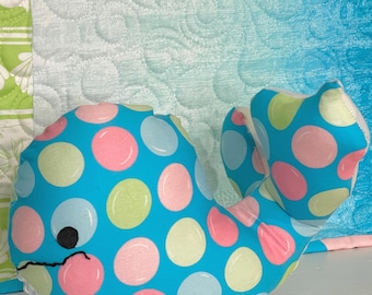 May include: A stuffed whale toy featuring a bright blue fabric base with a pattern of large, circular polka dots in shades of pink, light green, and pale blue. The whale has a simple black embroidered eye and a curved black stitched mouth.