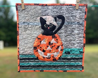 May include: A square quilted wall hanging featuring a black cat sitting inside a carved orange jack-o'-lantern. The background includes gray fabric with swirling patterns and text print, bordered by an orange fabric with black bat silhouettes.