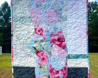May include: A rectangular fabric wall hanging featuring a quilted design of two ballet slippers. The slippers are decorated with a vibrant pink and blue floral pattern. The background includes sections of white fabric with a black line-art castle print.