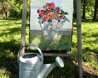 May include: A rectangular fabric wall hanging featuring a quilted design of a watering can filled with vibrant orange, red, and pink flowers, with a small bird perched on the spout, displayed above a matching galvanized metal watering can.
