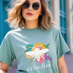 May include: A light teal t-shirt featuring a white bee design over a floral arrangement with the word "creative" in a script font. The shirt is worn by a person wearing sunglasses.