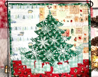 May include: A square quilted wall hanging featuring a central green and white gingham Christmas tree decorated with white snowflake appliques. The background includes panels of winter-themed fabric with cars, snowmen, and gifts in red and green.
