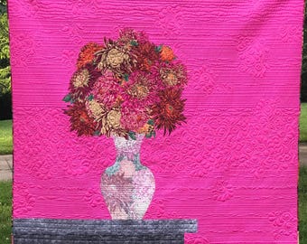 May include: A vibrant hot pink quilt featuring a central applique of a multi-colored floral bouquet in a patterned vase resting on a dark gray rectangular table. The fabric surface displays intricate horizontal line and swirl quilting patterns.