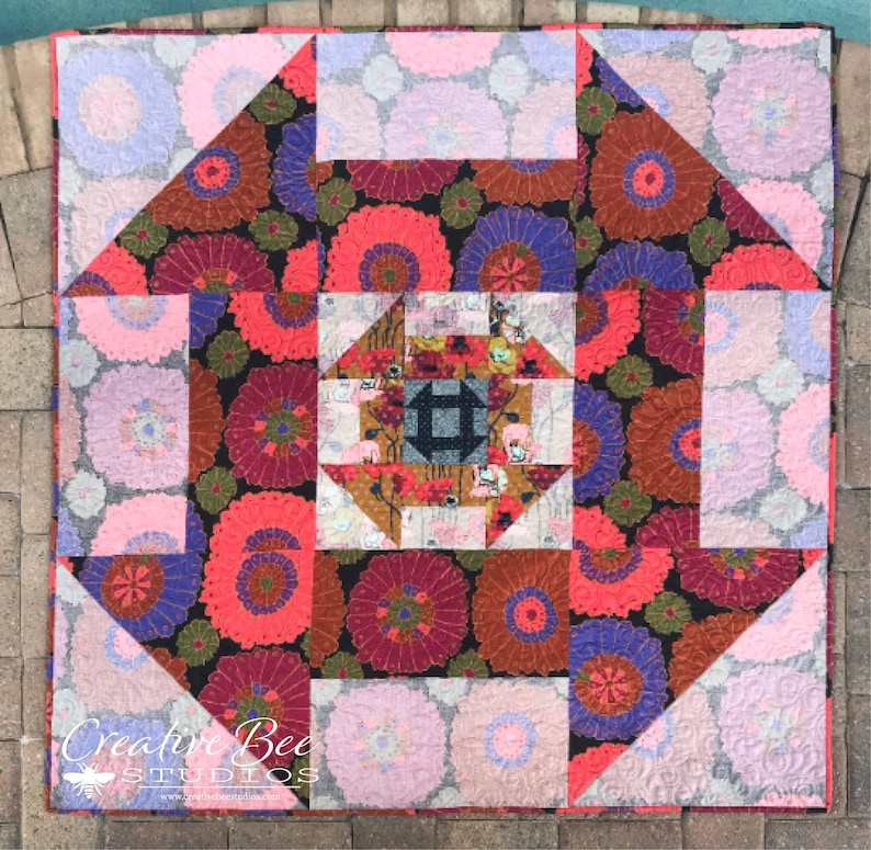 Op de afbeelding: Een quilt met een geometrisch patroon met een centraal vierkant met een kleiner vierkant erin. De quilt is gemaakt van verschillende stoffen met bloemmotieven in tinten roze, rood, paars en bruin. De quilt is omgeven door een rand van roze stof.