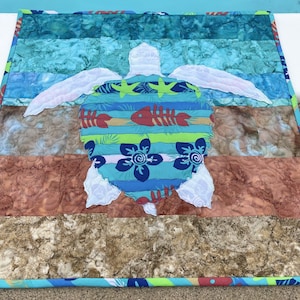 May include: A colorful patchwork quilt featuring a white sea turtle with a blue, green, and red patterned shell. The quilt is made up of various fabrics, including a blue and white striped fabric, a brown and white patterned fabric, and a red and white patterned fabric.