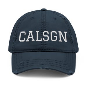 May include: A distressed navy blue baseball cap with the white text "CALSGN" across the front. The cap has a curved brim and a worn, vintage look.