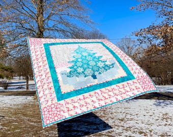 May include: A square quilt featuring a central teal Christmas tree graphic adorned with white snowflake patterns. The tree is framed by a solid teal border, surrounded by an outer patchwork border of pink and white circular and geometric motifs.