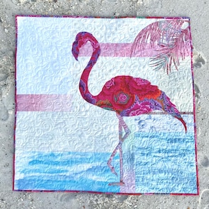May include: A square fabric quilt featuring a central flamingo applique in shades of hot pink, magenta, and purple with a floral pattern. The background displays a white sky with swirling quilted textures, a light pink horizon, and blue water.