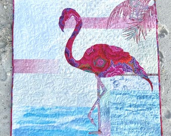 May include: A square fabric quilt featuring a central flamingo applique in shades of hot pink, magenta, and purple with a floral pattern. The background displays a white sky with swirling quilted textures, a light pink horizon, and blue water.