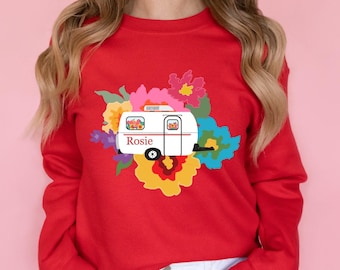 May include: A bright red crewneck sweatshirt featuring a central graphic of a white camper trailer with the name Rosie printed in red text. The camper is surrounded by a vibrant, multi-colored floral arrangement in shades of pink, yellow, and blue.